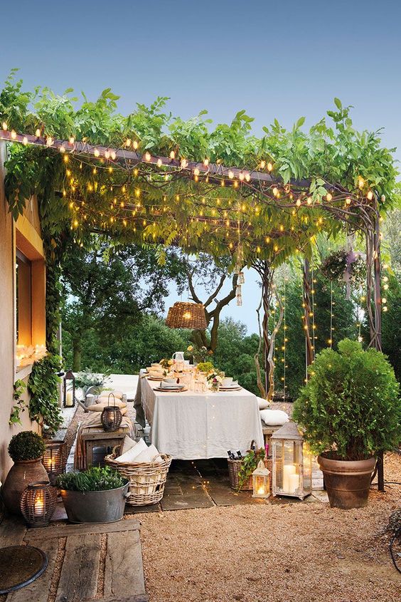 a lovely Provence terrace with wooden benches and a table, baskets with pillows, a woven pendant lamp and some lights over the space