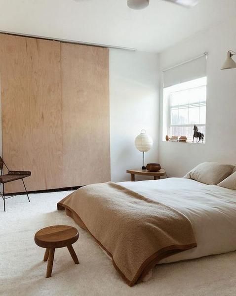 a natural and minimalist bedroom with a plywood storage unit, a bed with neutral bedding, wooden stools and a chair