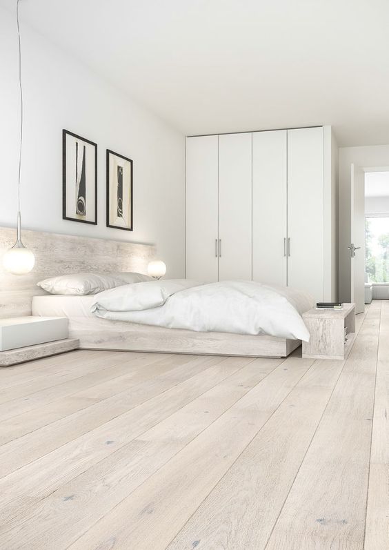 a neutral minimalist bedroom with a floor that matches the furniture, white bedding, a white wardrobe and a couple of artwork