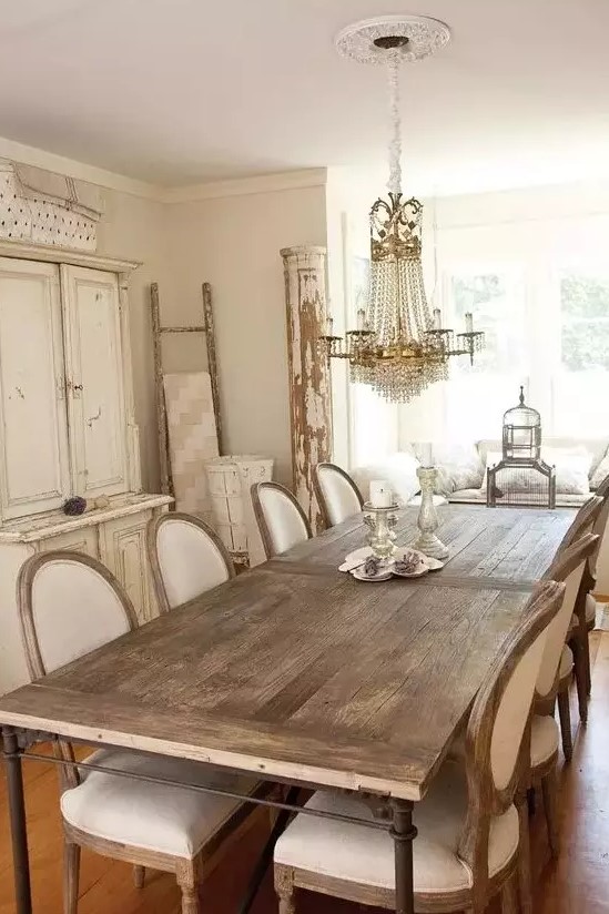 a neutral vintage dining room with a white storage unit, a stained dining table, neutral upholstered chairs, a crystal chandelier and some shabby chic decor