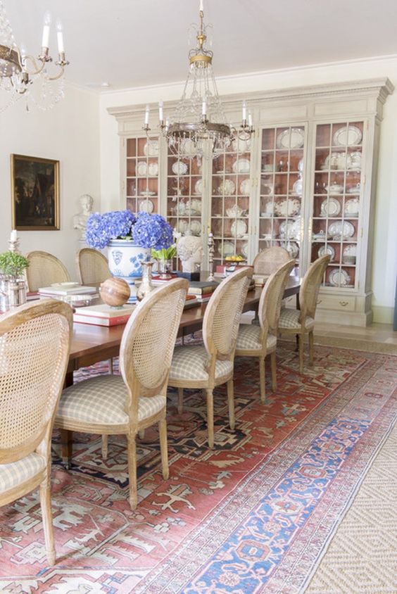a pretty French chic dining room with a neutral buffet with dishes, a stained table and neutral vintage chairs, layered rugs and crystal chandeliers