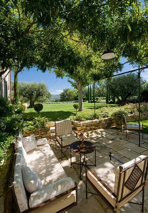 a pretty neutral Provencal terrace wiht neutral seating furniture, a round metal table, lots of greenery and trees around
