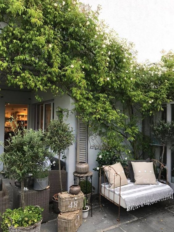 a small Provence-inspired terrace with a forged daybed with pillows, baskets and woven chests, potted greenery and trees