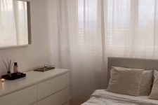 a small and cool minimalist bedroom done in neutrals, with a sleek white dresser, a pendant lamp, a creamy bed with neutral bedding, a mirror in a white frame