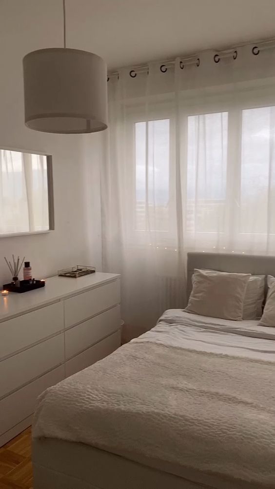 a small and cool minimalist bedroom done in neutrals, with a sleek white dresser, a pendant lamp, a creamy bed with neutral bedding, a mirror in a white frame