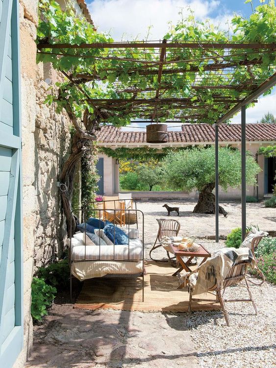 a small and lovely Provence outdoor space with a greenery roof, rattan chairs, a wooden coffee table, a metal daybed with printed upholstery