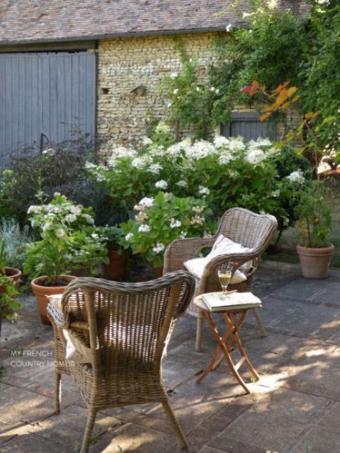 a small and lovely Provence terrace with woven chairs, a small side table, potted plants and greenery is a lovely nook to spend time in