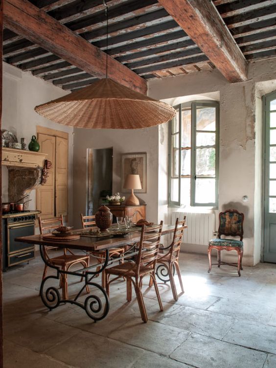 a vintage kitchen and dining space in one, with rich-stained wooden beams, a refined dining table and stained chairs, a wicker pendant lamp and some furniture
