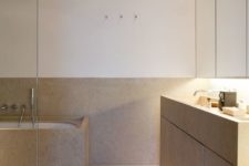 a warm-colored minimalist bathroom clad with tan-colored stone, a large vanity, a mirrored storage cabinet and a stone clad tub