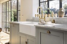 a beautiful dove grey farmhouse kitchen with shaker style cabinets, white stone countertops, a glazed wall and a large window with garden views