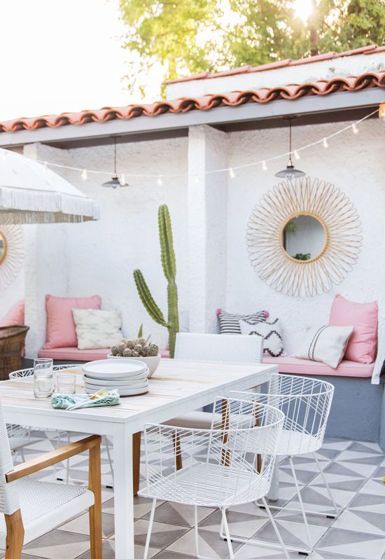 a beautiful modern patio with a tiled floor, a white dining table, white chairs, built-in benches with pink upholstery and a sunburst mirror