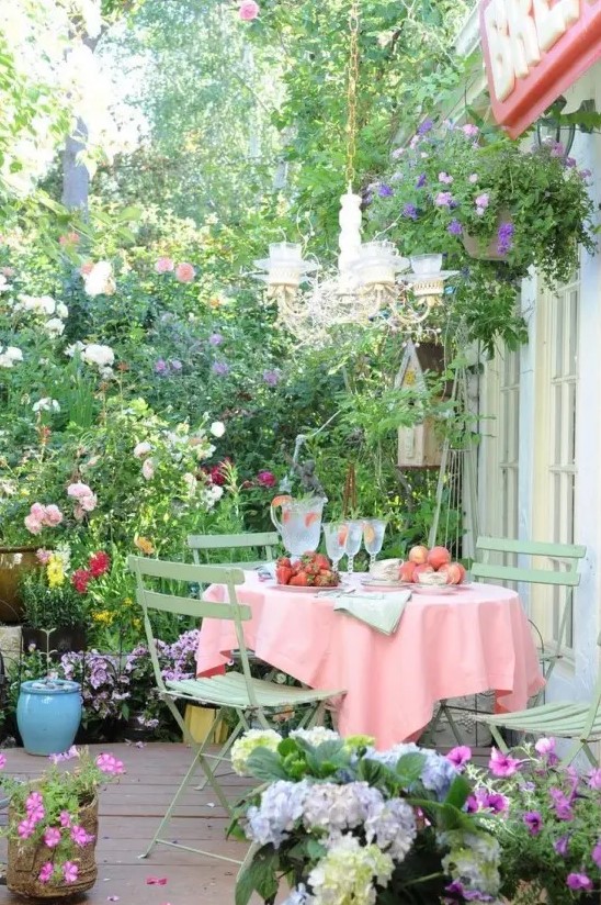 a bloom-filled spring terrace with potted greenery and flowers all around, with a dining set, a chandelier and some fresh fruit