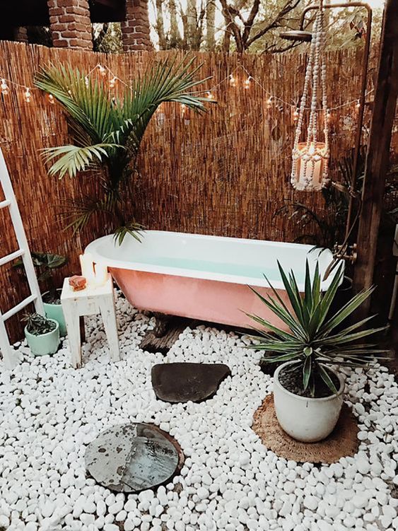 a boho chic outdoor space with tall woven walls, candles, pebbles, a pink bathtub and potted plants for a fresh feel