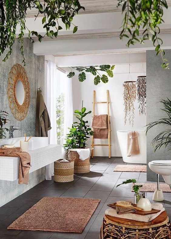a boho spa bathroom with a floating vanity, an oval tub, a rattan stool, a pink rug and towels, lots of potted greenery and greenery chandeliers
