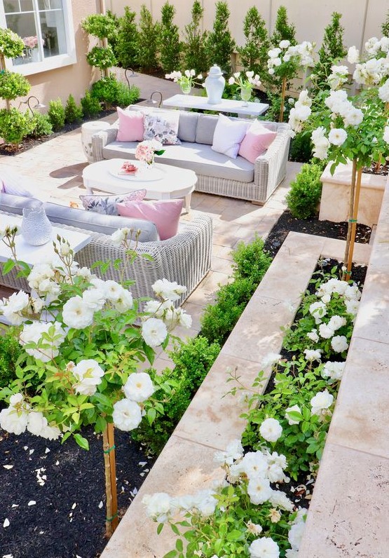a bright and vivacious summer terrace done with tiles, white wicker furniture, pastel upholstery and pillows and lots of greenery and blooms