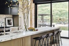 a chic light-stained and black kitchen with white marble countertops and black stools, a glazed wall with a mountain view