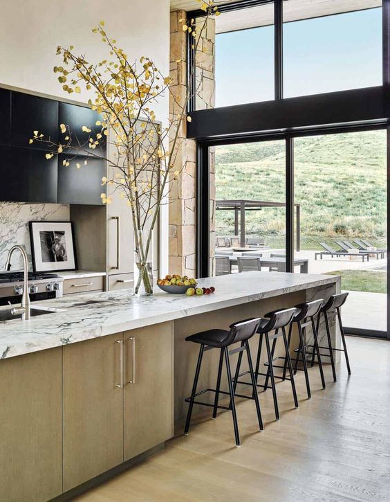a chic light stained and black kitchen with white marble countertops and black stools, a glazed wall with a mountain view