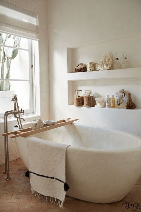 a chic neutral coastal spa bathroom with an oval stone tub, niches as shelves, wooden accessories and a window to the garden