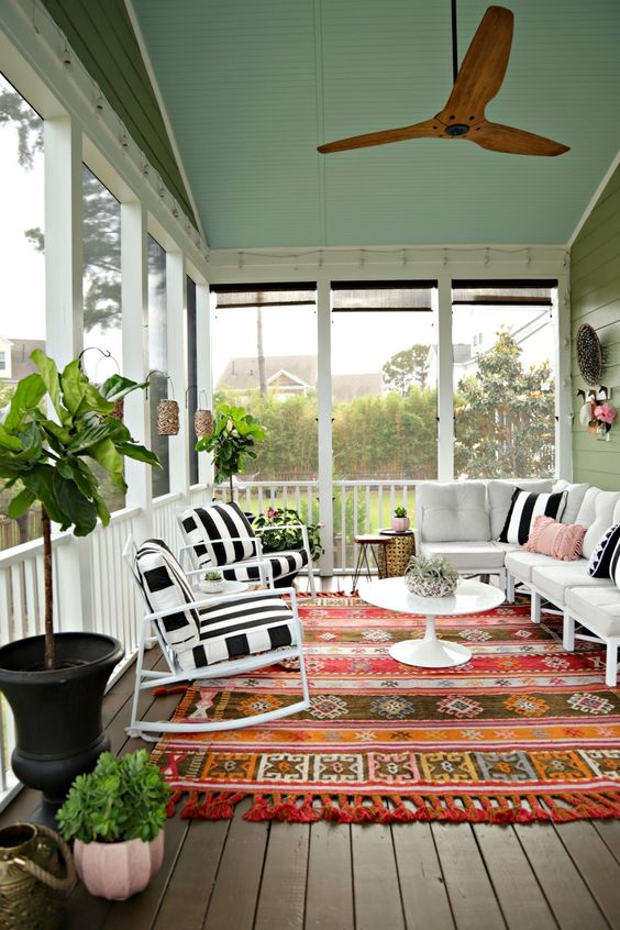 a colorful screened porch with white and black and white furniture, a colorful boho rug, potted plants and lanterns on the walls