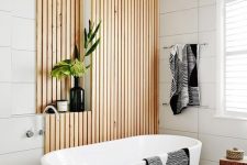 a contemporary neutral bathroom with white large scale tiles, wooden slats, a niche for storage, an oval tub and a wooden stool