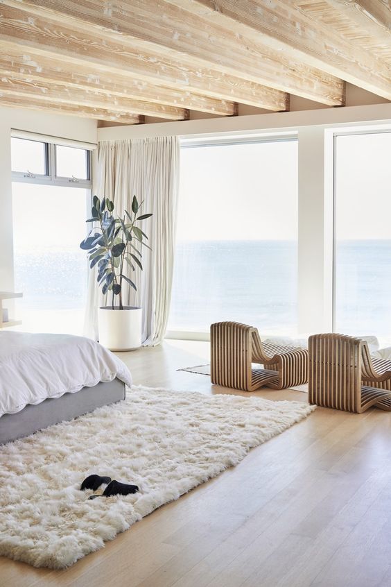 a cozy and stylish contemporary bedroom with catchy bent wooden loungers, a bed, a white fur rug and a statement plant plus sea views