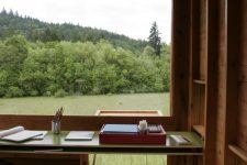 a cozy and welcoming modern home office clad with wood, with a glazed wall, a larrge desk and a chair plus views of the field and forests
