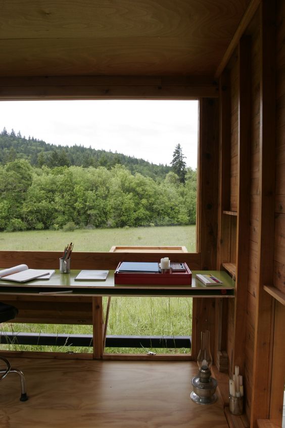 a cozy and welcoming modern home office clad with wood, with a glazed wall, a larrge desk and a chair plus views of the field and forests