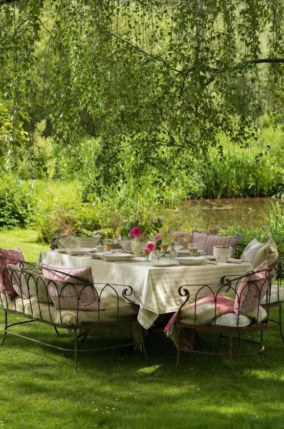 a delightful Provence outdoor space with a dining set of metal, with lots of cushions and pillows, bright blooms and greenery over it