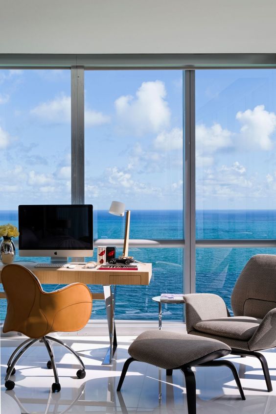 a fab home office with a glazed wall and a lovely sea view, a desk, an amber chair, a grey chair to relax in and a footrest