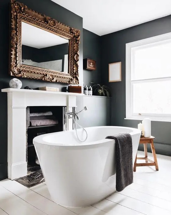a fancy bathroom with black walls and a white planked floor, a fireplace used for storing towels, an oval tub and a mirror in a refined frame