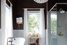 a glam bathroom with chocolate walls, white paneling, a vintage tub, a chic crystal chandelier and vintage furniture just wows