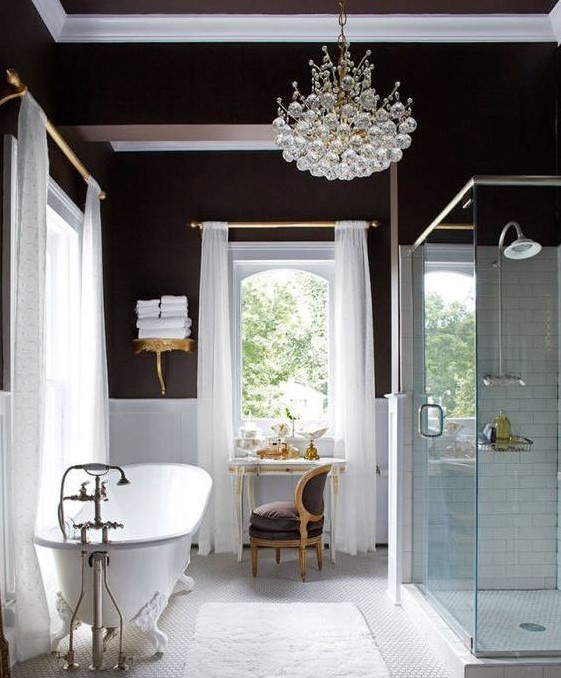 a glam bathroom with chocolate walls, white paneling, a vintage tub, a chic crystal chandelier and vintage furniture just wows