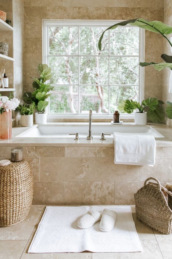 a gorgeous neutral spa bathroom clad with limestone tiles, with a tub clad with them, built in shelves and potted greenery, a woven pouf and a bag