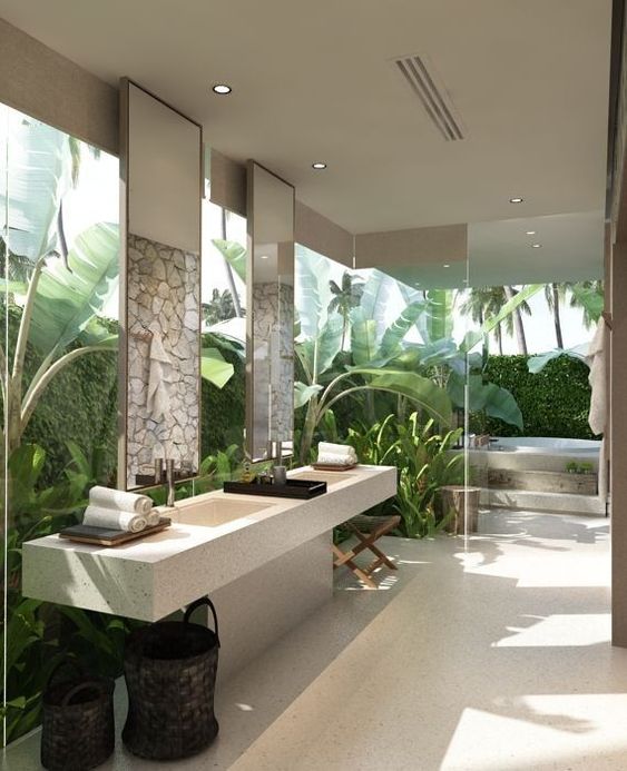 a jaw dropping tropical spa bathroom with glass walls and surrounded with greenery and tropical plants, a tub, wooden stools, a stone vanity and some neutral towels