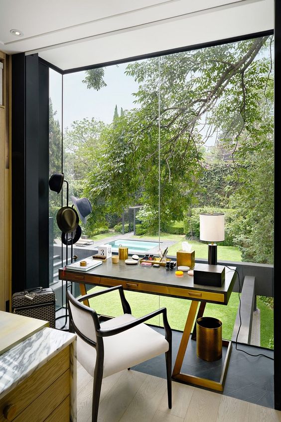 a lovely and cozy home office nook with a glazed wall and a view of the garden, a chic desk, a creamy chair and some brass decor
