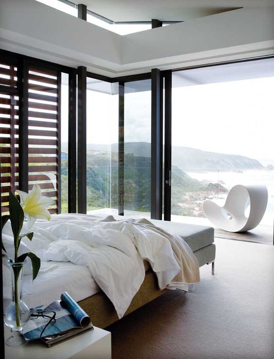 a lovely contemporary bedroom with a bed, a bench, nightstands, glass walls and a balcony plus amazing ocean views