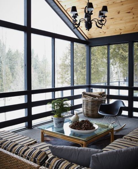a lovely modern screened porch with a striped corner sofa, a rattan coffee table, a black chair and a basket with firewood