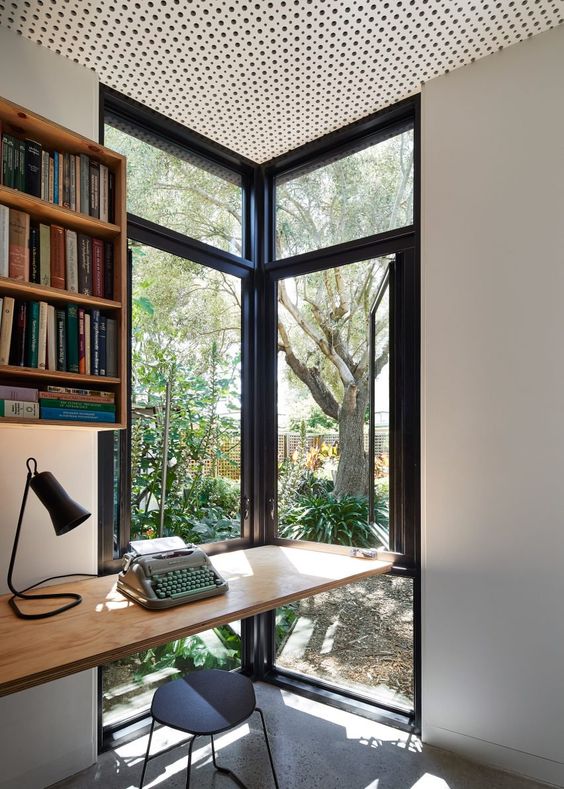 a lovely small home office nook with a corner window that lets enjoy garden views, a floating desk, a black stool and a shelving unit