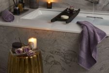 a luxurious spa bathroom clad with grey marble tiles, a tub, a gold side table, candles, scents and some blooms