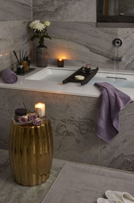 a luxurious spa bathroom clad with grey marble tiles, a tub, a gold side table, candles, scents and some blooms