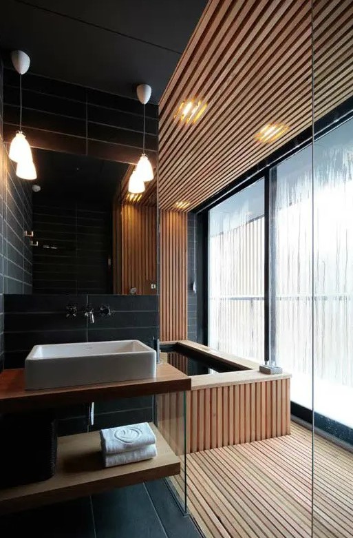 a minimalist bathroom done with light stained wooden slab and black skinny tiles, with pendant lamps and a large window for more light