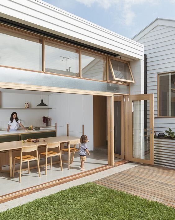 a minimalist kitchen with white cabinets, a dining zone with stained furniture, a glass wall that can be removed