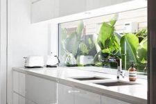 a minimalist sleek white kitchen with neutral stone countertops, a a window backsplash with a tropical garden view