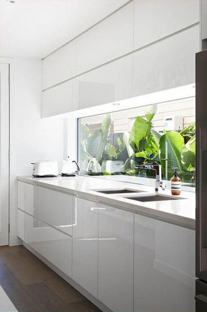 a minimalist sleek white kitchen with neutral stone countertops, a a window backsplash with a tropical garden view