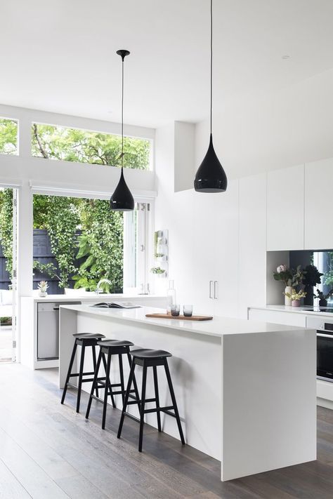 a minimalist white kitchen with sleek cabinets, white stone countertops, black pendant lamps and a glazed wall that looks on the garden