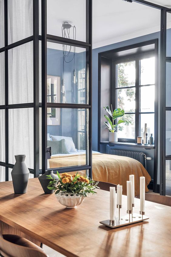 a modern bedroom with blue walls, a bed, some statement plants and vases and glass walls all over to maximize the daylight