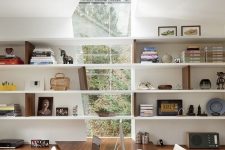 a modern home office with a catchy irregular window to enjoy garden views, lots of wall-mounted shelves, sleek storage units and a built-in desk