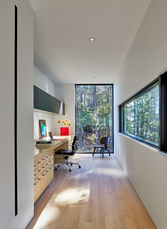 a modern home office with a glazed wall and a long and narrow window, a large desk, black chairs, a sleek storage unit and amazing views of the forest