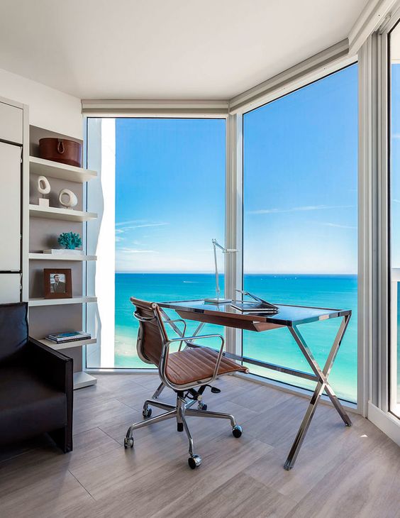 a modern home office with glazed walls and a gorgeous sea view, a modern trestle desk, a brown leather chair, built-in shelves and a black sofa