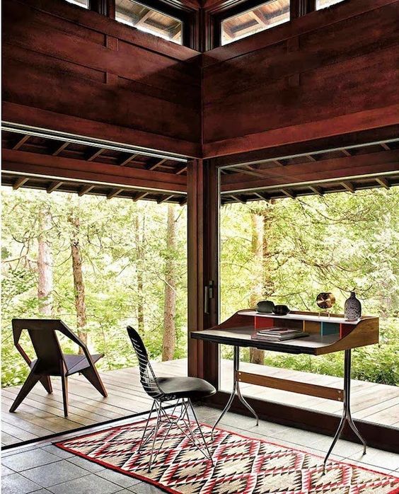 a modern home office with glazed walls, stained wood and a stained desk, cool chairs and a printed rug plus views of the forest